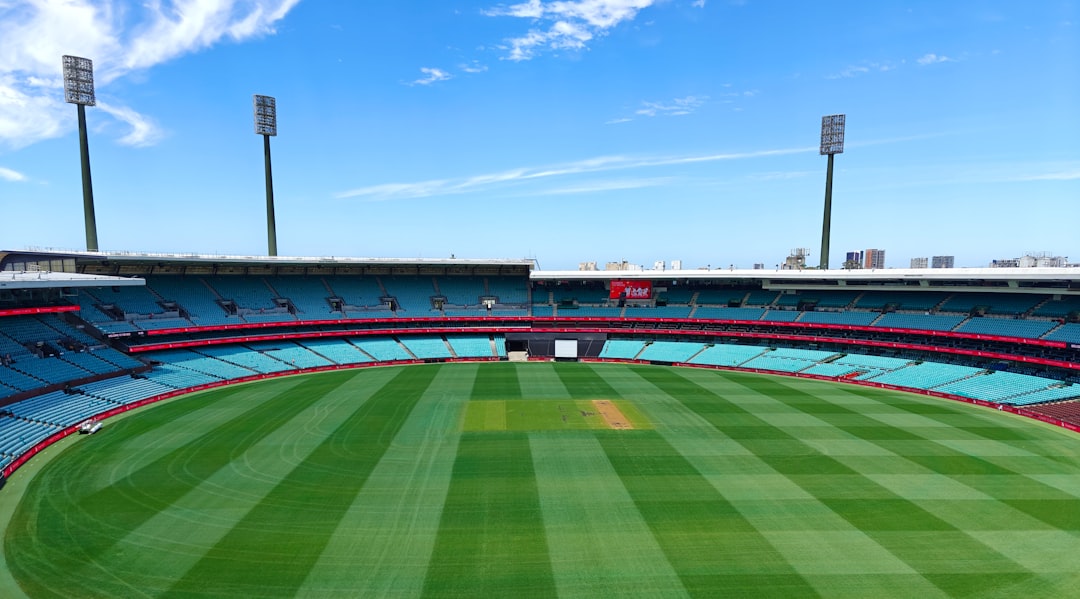 Wide-angle view of a cricket stadium featuring a green pitch and empty seating under a blue sky.